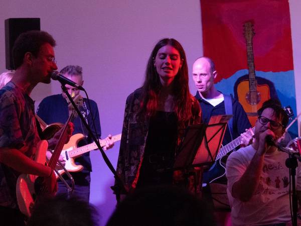 Sängerin vor Mikrofon, Links ein Gitarrenspieler, rechts ein weiterer Sänger sitzend, im Hintergrund zwei Bandmitglieder