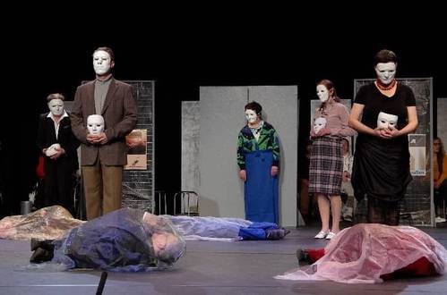 Menschen stehen mit weißen Masken auf der Bühne, davor liegen zwei Menschen zugedeckt mit Tüchern.