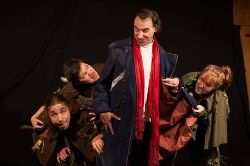 Vier Personen in Kostümen, eine in der Mitte mit rotem Schal, wirken theatralisch.