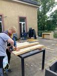 Zwei Personen sind auf einer Terrasse damit beschäftigt, Holzbretter zu verarbeiten. Eine Person benutzt einen Akkuschrauber, während die andere die Holzbretter hält.