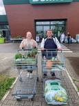 Zwei Personen stehen vor einem Baumarkt und halten jeweils einen Einkaufswagen voller Pflanzen und Gartenartikel. Vor ihnen liegen verschiedene Kräuter, Blumen und ein großer Sack Blumenerde.